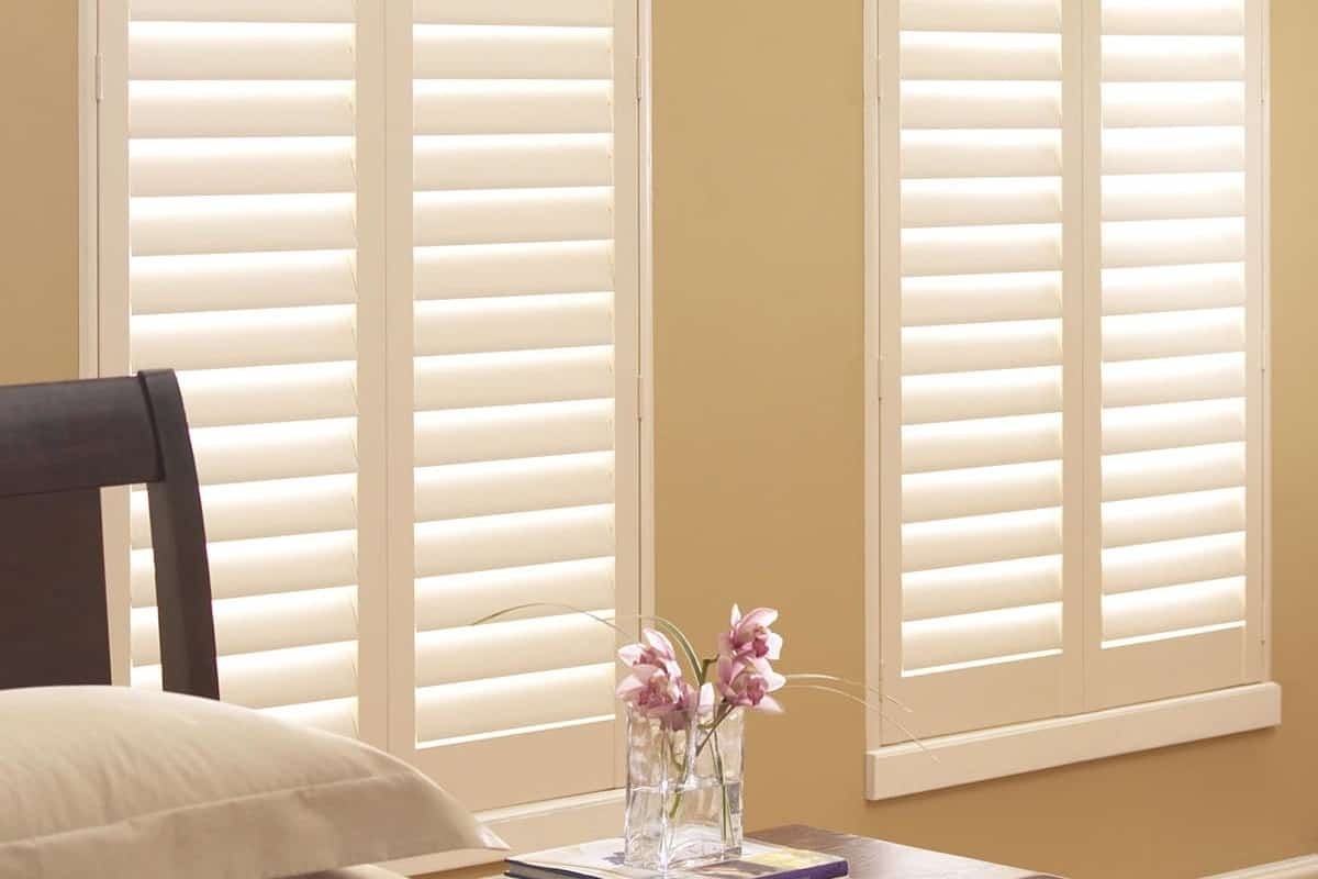 Closed white Palm Beach™ Polysatin™ Shutters hanging over a comfy-looking bed and side table